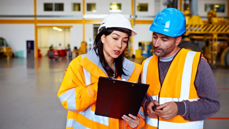 two warehouse workers talking together over a clipboard while standing inside of a large warehouse